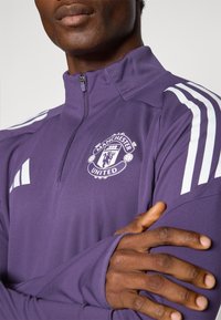 Purple athletic zip-up jacket with white stripes on shoulders and a Manchester United logo on the chest. Smooth fabric, fitted design.