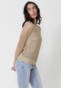 Beige kötött pulcsi laza, nyitott szövésű kialakítással. Hosszú ujjakkal és kerek nyakkivágással rendelkezik, világoskék farmerral párosítva.