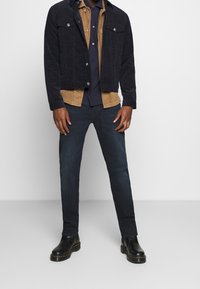 Mörkblå kordjacka med tryckknappar över en beige kordskjorta, i kombination med mörka jeans och svarta läderankelboots.