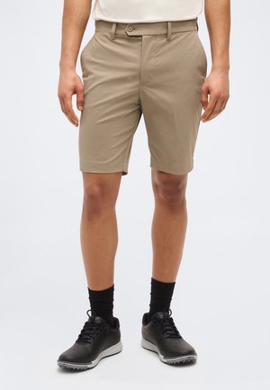 Uomo con pantaloncini beige fino al ginocchio, calzini neri e scarpe nere stringate, in piedi contro uno sfondo chiaro e semplice.