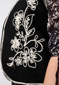 Zwart suede kledingstuk met witte bloembroderie, decoratieve draad en parelaccenten. Lange mouwen met een gedetailleerde stofpatroon.