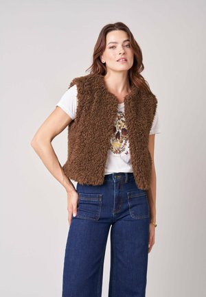 Vrouw die een bruine pluizige vest draagt over een wit t-shirt met print en donkerblauwe hooggetailleerde spijkerbroek met voorzakken.