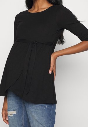 Femme portant un haut de maternité noir à manches longues avec un lien sous la poitrine et un jean bleu déchiré.