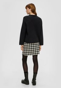 Zwarte trui met lange mouwen, gecombineerd met een rok van hounds-tooth patroon in crèmewit en zwart. Model draagt zwarte panty’s en stevige zwarte laarzen.