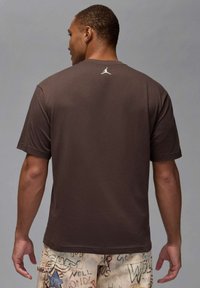 Camiseta marrón de manga corta, cuello redondo, material de algodón, con un logo blanco en la parte superior trasera, combinada con pantalones cortos claros estampados.