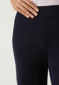 Mörkblå stickade byxor med en mjuk textur, med ett brett midjeband och sömdetaljer på sidan, visade med en hand som vilar på tyget.