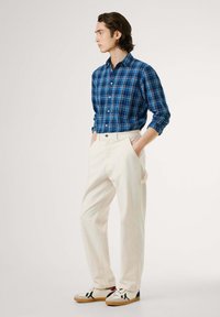 Blauwe geruite knopshirt, crèmekleurige broek, witte sneakers met zwarte en rode accenten. Alleenstaand, zijaanzicht tegen een neutrale achtergrond.