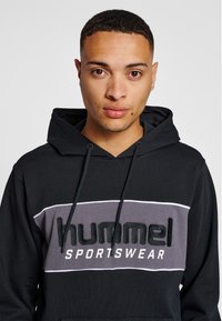 Svart hoodie med en grå och vit horisontell panel. "Hummel"-logotyp i svart velour på bröstet. Justerbar huva med dragsko.