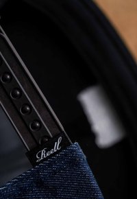Una tela de mezclilla de un tono azul oscuro presenta una pequeña etiqueta negra que dice "Reell". El hardware metálico con puntos en relieve y un diseño elegante son visibles.