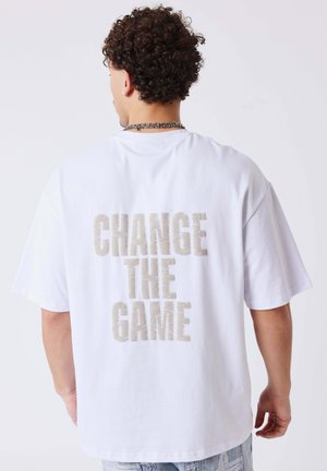 Man met krullend haar, draagt een wit T-shirt met de tekst "CHANGE THE GAME" op de achterkant, een zilveren ketting en een lichtblauwe spijkerbroek.