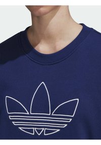 Detailní záběr osoby v námořnickém modrém svetru s bílým logem Adidas trefoil na hrudi.