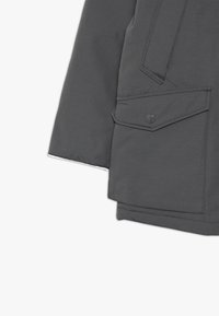 Manche de veste gris foncé avec une poche à rabat sécurisée par un bouton-pression, montrant des coutures détaillées sur un tissu lisse.