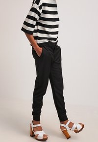 Przerysowany czarno-biały top w paski, czarne zwężane spodnie oraz białe sandały na drewnianych obcasach, prezentujące nowoczesny, swobodny styl.