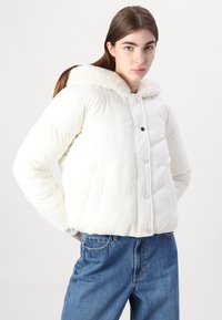 White puffer jacket with a plush, fur-lined hood, front snap buttons, and side pockets, paired with blue jeans. Smooth, quilted texture.
