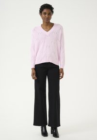 Pull-over en V rose en tricot avec un motif en losanges texturé. Associé à un jean noir à jambes larges et des bottines noires à finition lisse.