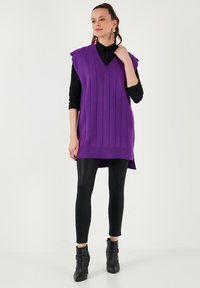 Purple knitted sleeveless vest with vertical ribbed texture, V-neck design, paired with black leggings and ankle boots.