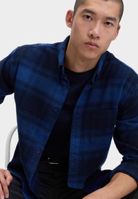 Młody mężczyzna z krótkimi włosami, ubrany w niebieską i czarną koszulę w kratę z guzikami, na czarnej koszulce, siedzący na białym krześle.