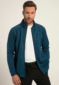 Blaue Zip-Jacke aus strukturiertem Stoff, hoher Kragen und zwei Seitentaschen. Mit schwarzen Akzenten entlang des Reißverschlusses und an den Bündchen.