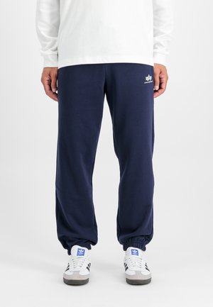 Pantalones de chándal azul marino con puños elásticos, hechos de una tela suave, que presentan un pequeño logo blanco en el muslo izquierdo. Llevados con zapatillas blancas.