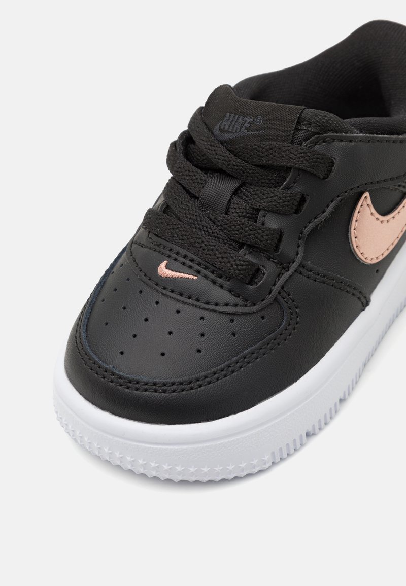 Baskets en cuir noir avec semelle en caoutchouc blanche, présentant un swoosh Nike rose et des lacets noirs. Surface texturée avec des perforations à l'orteil.