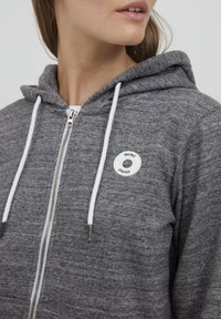 Sivi zip-up dukserica s bijelim vezicama i okruglim bijelim logotipom na prsima. Tkanina izgleda mekano s blagom teksturiranom površinom.