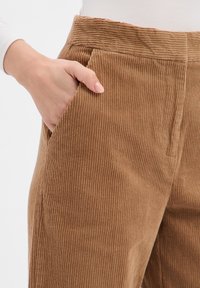 Brązowe sztruksowe spodnie w pionowe paski, z boczną kieszenią i miękką teksturą. Pas jest lekko podniesiony.