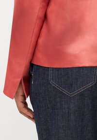 Blazer di raso corallo con maniche lunghe, fessure laterali e una finitura liscia, abbinato a jeans in denim scuro con un design classico delle tasche.