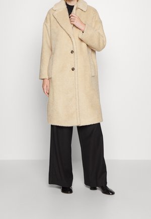Personne portant un manteau beige duveteux longueur genoux avec un col et deux boutons, associé à un pantalon large noir et des chaussures noires.