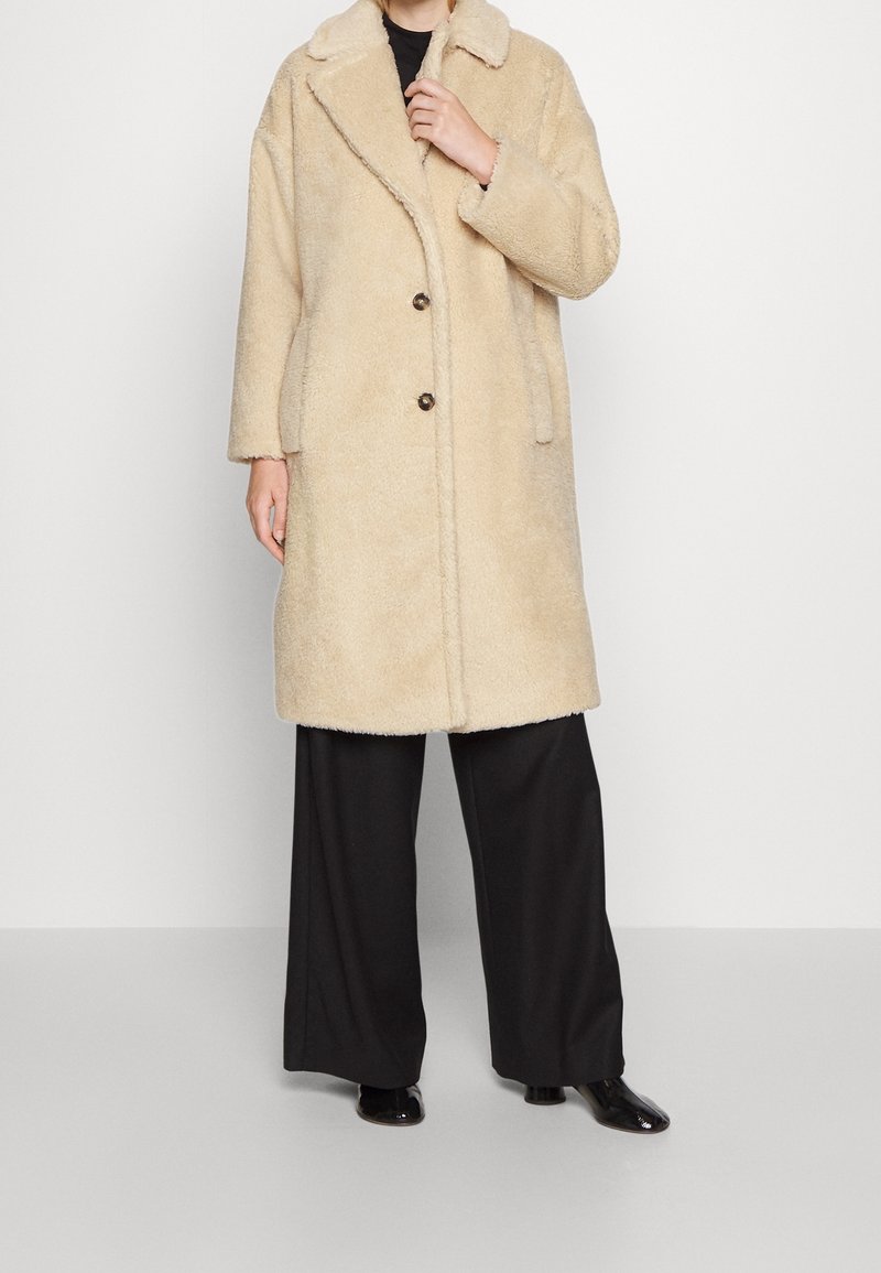Personne portant un manteau beige duveteux longueur genoux avec un col et deux boutons, associé à un pantalon large noir et des chaussures noires.