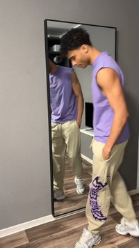 Νεαρός άνδρας με μωβ αμάνικο πουκάμισο και μπεζ παντελόνι με μωβ σχέδιο κοιτάζει κάτω, όρθιος δίπλα σε έναν καθρέφτη ολόσωμο σε ένα δωμάτιο.