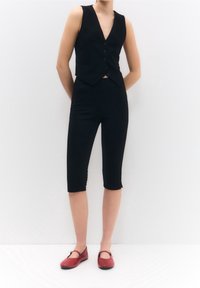 Gilet nero senza maniche con chiusura a cinque bottoni, abbinato a pantaloni corti neri aderenti fino al ginocchio e ballerine rosse. Tessuto liscio, design minimale.