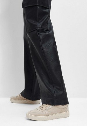 Pantaloni in pelle nera con una texture liscia e design a gamba larga, abbinati a sneakers in suede beige con dettaglio zip e suola a righe.