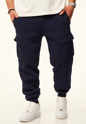 Mann trägt marineblaue Cargo-Jogger-Hose mit großen Seitentaschen und weißen Sneakers, Hände in den Taschen, steht vor schlichtem Hintergrund.
