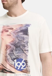 Witte katoenen T-shirt met een grafische print van een abstract strandtaferelen in paarse en beige tinten. Circulair logo in het blauw met witte tekst.