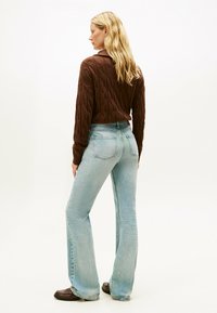 Brązowy sweter w splot kablowy zestawiony z jasnoniebieskimi dżinsami w kształcie dzwonów. Dżinsy mają średni wzrost w talii i lekkie wyblaknięcia. Widać płaskie brązowe buty.