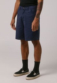 Tamsiai mėlyni medvilniniai šortai su lygiu priekiu, šoninėmis kišenėmis ir mygtuku, derinami su juodais slip-on tipo bateliais ir juodomis banguotomis kojinėmis.