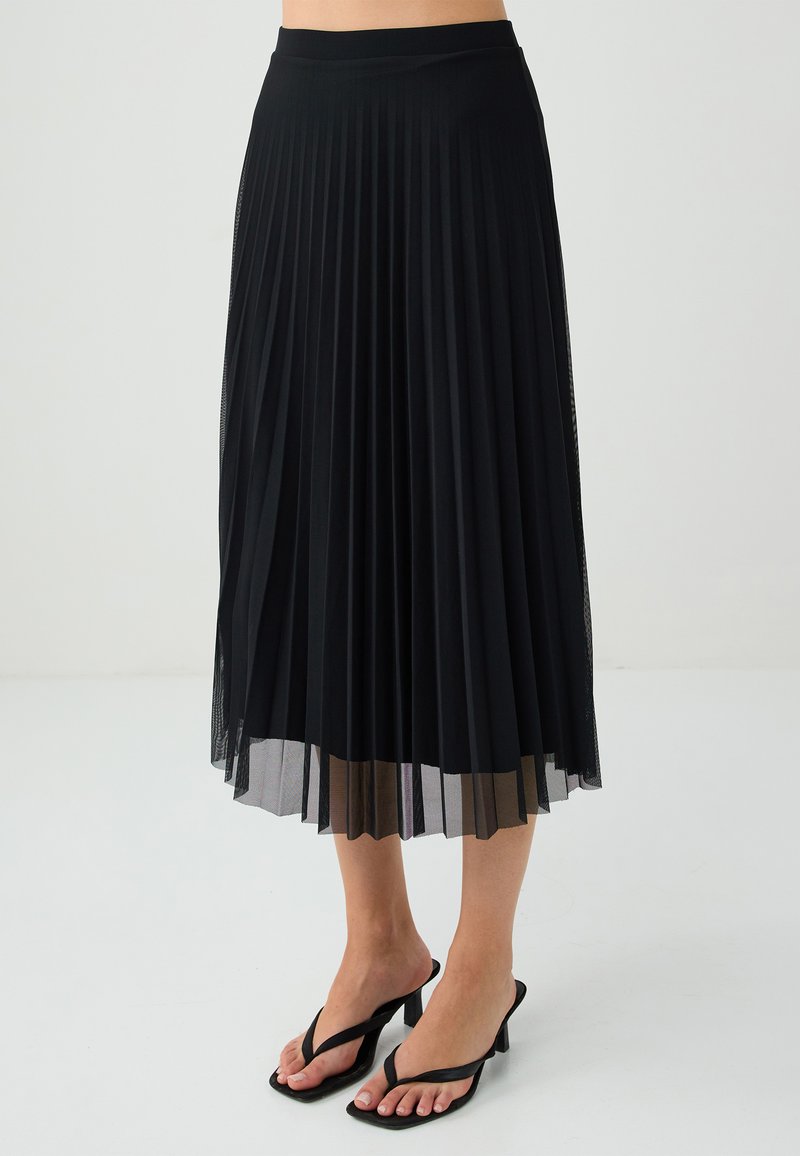 Schwarzer plissierter Midirock mit glattem Bund und transparenter unterer Schicht. Kombiniert mit schwarzen Riemchenabsätzen, die ein minimalistisches Design präsentieren.