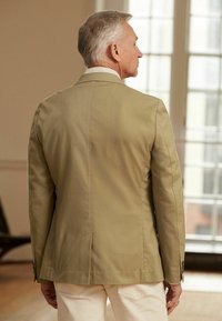 Kaki blazer med et struktureret design, enkelt ventil på bagsiden og to knapper på ærmerne. Bæres over en lys skjorte.