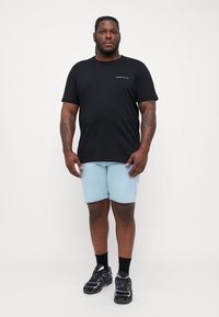 Sort bomulds t-shirt med grønne trykte bogstaver, kombineret med lyseblå denimshorts. Modellen bærer sorte sneakers og sokker.
