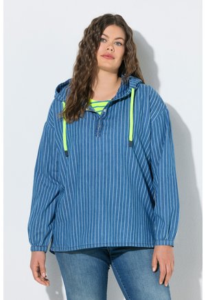 Frau mit langem welligem Haar trägt einen blauen Kapuzenpullover mit Nadelstreifen und neongelben Kordeln über einem neongrün gestreiften Oberteil und blauen Jeans.