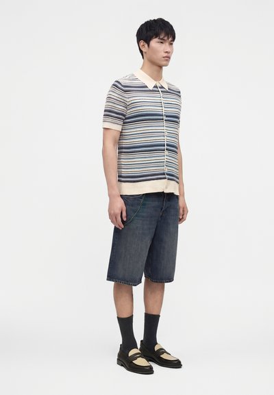 Jeune homme portant une chemise à manches courtes rayée boutonnée, un short en jean au genou, des chaussettes noires et des mocassins bicolores, debout sur un fond blanc.