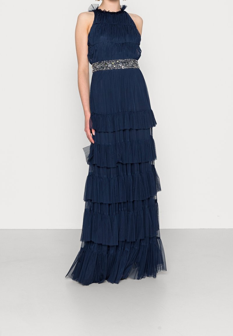 Marinblå maxiklänning med flera lager och volangdetaljer, prydd med bälte. Lättviktsmaterial med en flödande design och genomskinliga inslag.