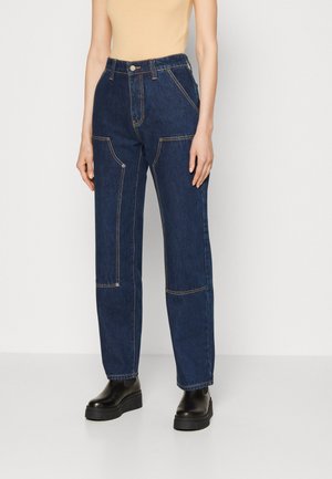 Femme portant un jean denim bleu foncé taille haute avec coutures contrastées, poches avant, et des bottes plateformes noires épaisses, debout devant un fond blanc.