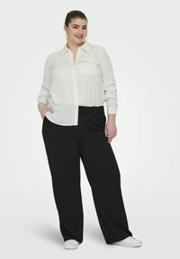 Cămașă albă cu mâneci lungi, cu buzunar pe piept, și pantaloni negri cu croială largă, cu bucle pentru curea; purtată cu adidași albi.