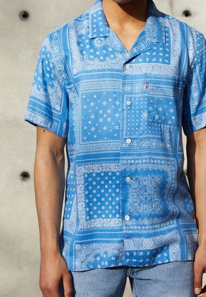 Hombre con camisa azul de manga corta con botones y estampado blanco estilo bandana y vaqueros azul claro contra un fondo gris.