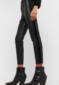 Zwarte nepleren broek met een nauwsluitende pasvorm, voorzien van verticale zwarte fluwelen strepen langs de zijkanten en metalen knoopaccenten.