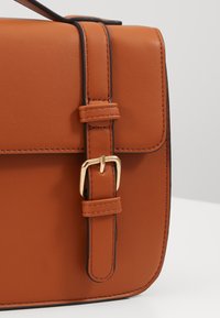 Sac à main en cuir marron avec une texture lisse, présentant un détail de boucle en or, des surpiqûres noires et une forme structurée.