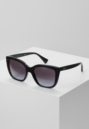 Lunettes de soleil noires surdimensionnées avec des verres dégradés, des montures en plastique épaisses et des détails de logo sur la branche. Exposées sur un piédestal blanc.