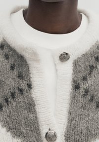 Cardigan gris et blanc en tricot avec une texture duveteuse, doté d'un bouton argenté et d'un col blanc contrastant sur un vêtement blanc.