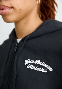 Crna zip jakna od mekog materijala s bijelim logom "New Balance Athletics" na lijevoj strani prsa. Glatka tekstura i ležeran dizajn.
